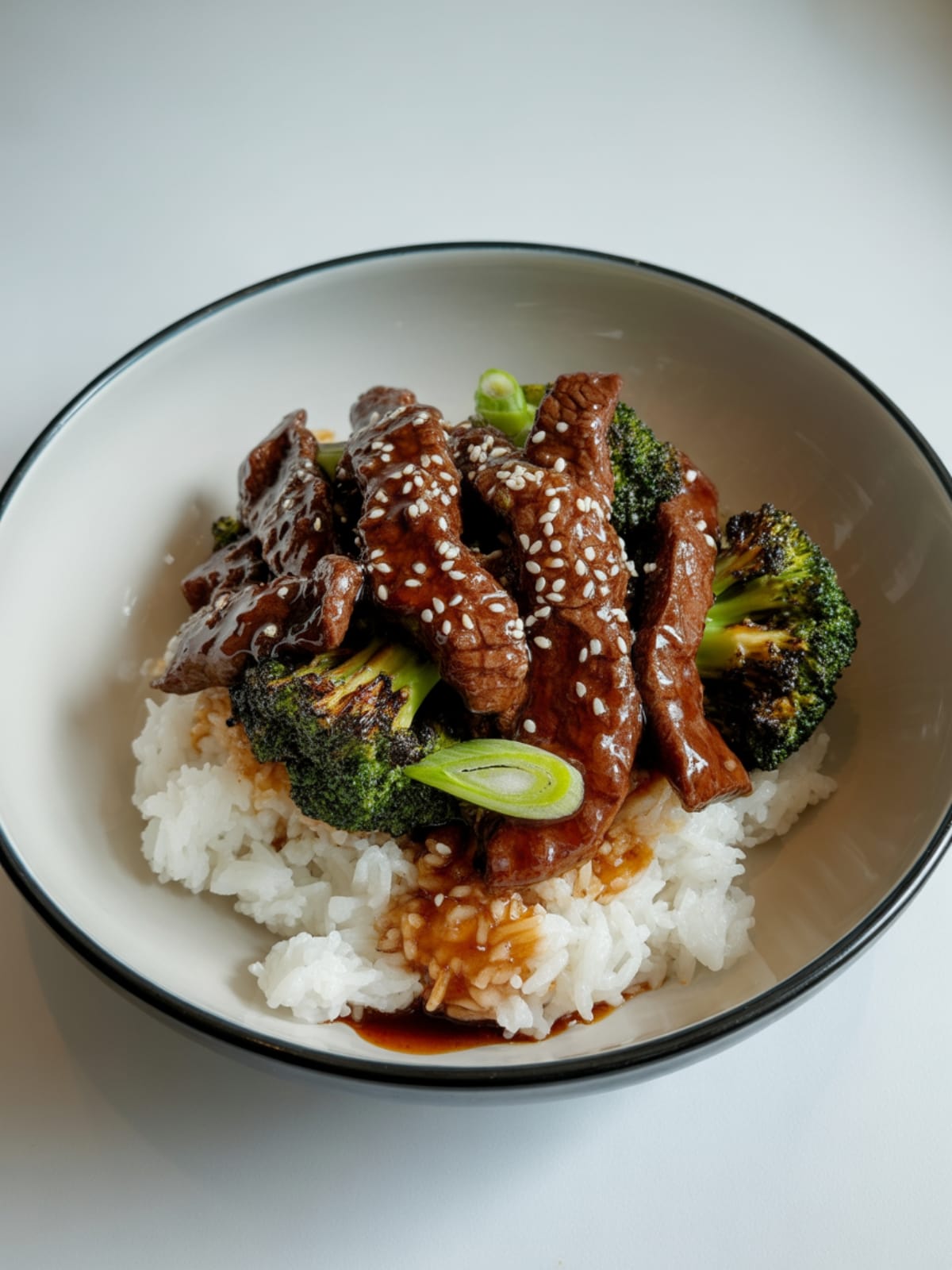Beef and broccoli stir-fry in Asian bowl over rice, garnished with scallions and sesame seeds